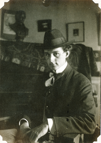 Ralph Warner, looks almost temptingly at the camera, chin tilted down, hat on while sitting in a chair in a haphazardly decorated room.