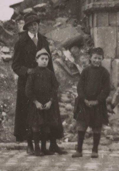     Photograph shows American Red Cross representative for the district of Attigny, Miss Edna M. Walker of Woodstock, New York, standing with two French refugee orphaned children in front of the ruins of the church in Attigny, France. The caption on verso indicates that Charlemagne had passed through the arch in 825, although the French emperor died in 814, and the Roman-style Église Notre-Dame d'Attigny was constructed centuries after his reign.