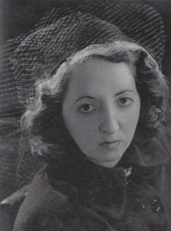 A young woman looks into the camera with a serious expression, her short, wavy hair topped by a hat with veiling.