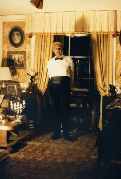 Ted Pierce poses in a living room near a window. He is wearing a tuxedo, with no jacket.
