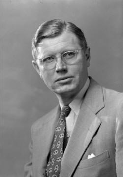 Close-up of a man in a suit with a patterned tie and clear-rimmed glasses.